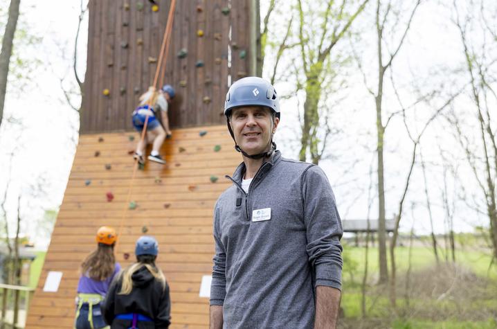 Roger Brown standing in front of an outdoor climbing wall as students climb behind him