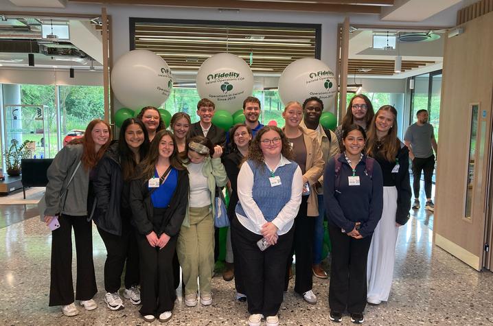 A large group of people stand together in a bright indoor lobby, posing for a group photo beneath three large white balloons, with windows and a hallway in the background.