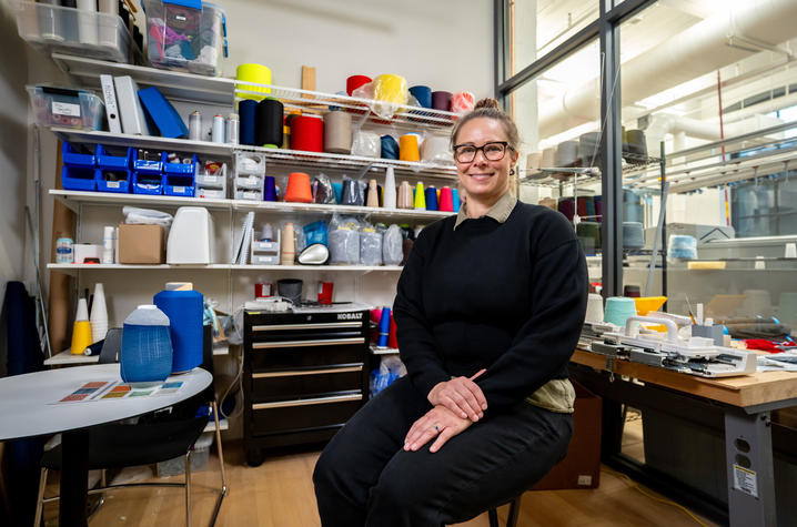 A person in a black sweater sits on a stool in a textile workshop, hands folded in their lap. 