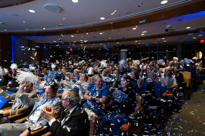 A large group of people seated in an auditorium, wearing blue and white attire and accessories such as scarves and pom-poms. Blue and white confetti fills the air, creating a festive atmosphere. 