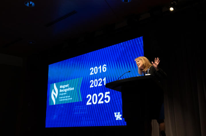 A woman stands at a podium on a stage, gesturing with one hand raised, in front of a large screen displaying the text “Magnet Recognition” along with the years “2016,” “2021,” and “2025.”