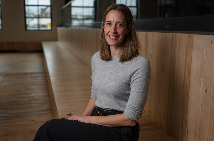 photo of Kristi Bartlett A person with shoulder‑length brown hair sits on wooden bleacher-style seating in a modern, spacious room with large industrial-style windows. They are wearing a light gray long-sleeve top and a dark skirt or pants, with hands resting in their lap.