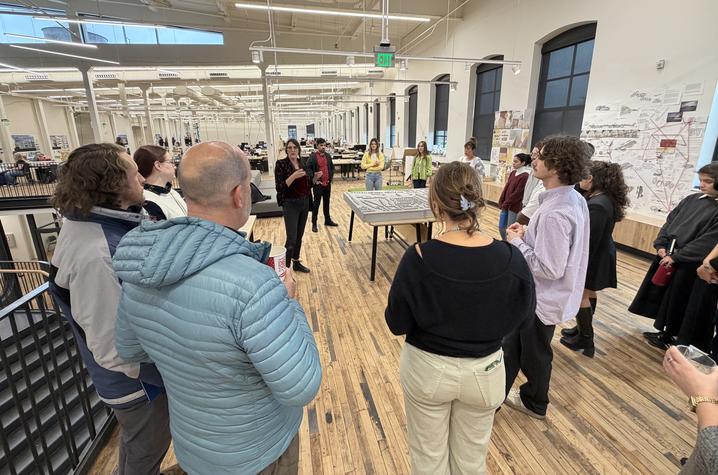 A group of people stands in a spacious, modern office or studio with high ceilings, wooden floors, and large windows. They appear to be listening to someone speaking near the center of the room