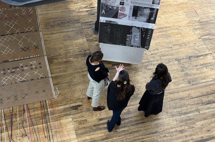 An overhead view of three people standing on a worn wooden floor, gathered around a large vertical display board with architectural drawings or design plans. One person gestures with their hand while the others listen. 