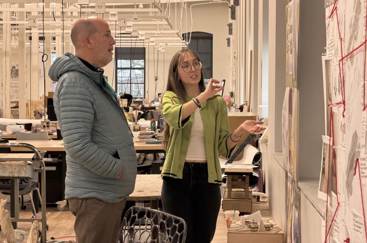 A man and a woman stand in a bright, open studio space, looking at a large wall display covered with sketches and diagrams. The woman gestures toward the artwork while the man listens.