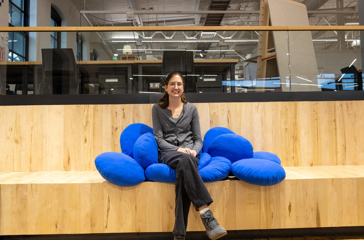 A woman wearing glasses and a gray cardigan sits smiling on a bright blue cushioned bench inside a modern building with wood paneling, large windows and exposed industrial-style ceilings in the background.