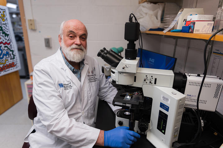 A researcher in a white lab coat and blue gloves sits beside a large microscope, facing the camera. Lab shelves, boxes and instruments surround the workstation.