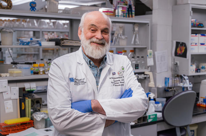 An older researcher in a white lab coat and blue gloves stands with arms crossed in a laboratory. Benches, bottles, racks and lab equipment fill the background.