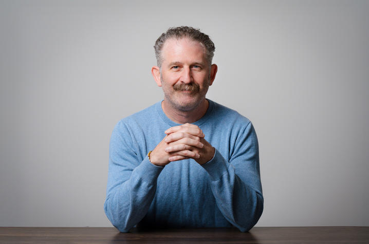 photo of Ashley Seifert Studio portrait of an adult with short curly hair and a mustache, seated at a table with hands clasped. The person wears a light blue sweater against a plain light gray background.