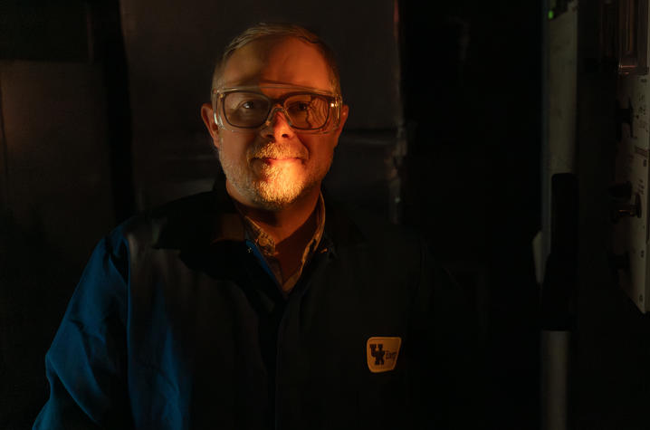 A close-up portrait of a person wearing safety glasses, lit by a warm light from the side in a dark laboratory setting.