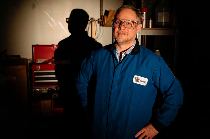 Matt Weisenberger in a blue UK Energy lab coat and safety glasses, standing in a dimly lit workshop with a red toolbox and equipment in the background.
