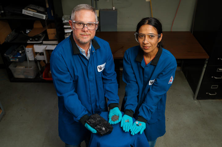 Two people in blue lab coats and safety glasses, holding a piece of coal, powdered coal and finished graphite discs.