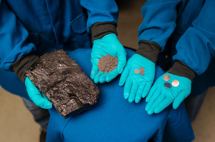 Hands in teal gloves hold a large piece of coal, a pile of grey powdered coal and several small metallic graphite discs against a blue background.
