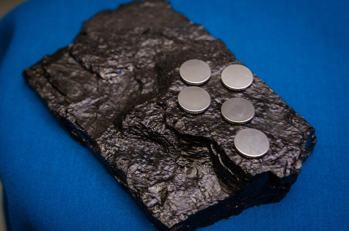 A large, jagged piece of black coal resting on blue fabric, with five small, shiny metallic graphite discs placed on its surface.
