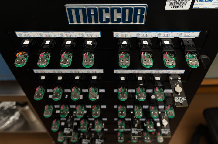 Overhead view of a black MACCOR battery testing units with numerous orange and black wires ending in alligator clips attached to small battery samples.