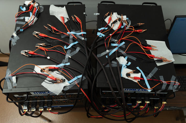 Overhead view of two black MACCOR battery testing units with numerous orange and black wires ending in alligator clips attached to small battery samples.