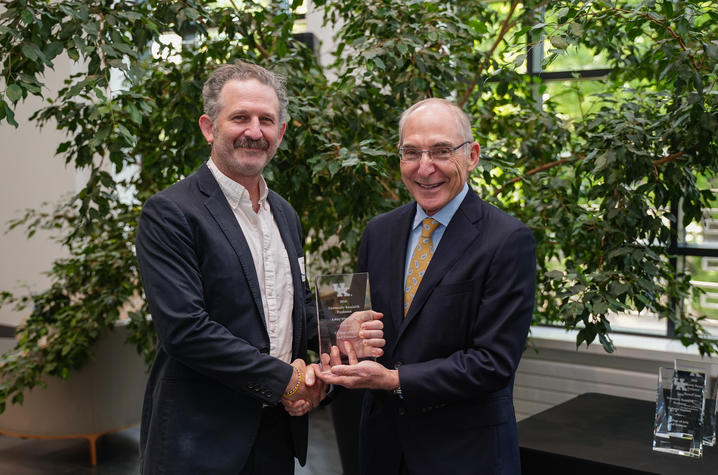 photo of Seifert and Capilouto Two people stand indoors among large leafy plants, shaking hands while holding a glass award. The recognition photo captures an academic honors presentation in a bright atrium-like setting.