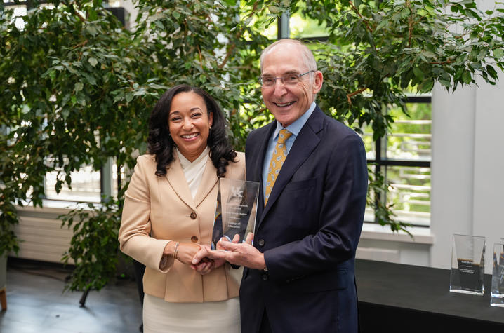 Two people smile and shake hands while holding a glass award engraved “University Research Professor.” They stand in a bright indoor space with leafy plants and large windows in the background.