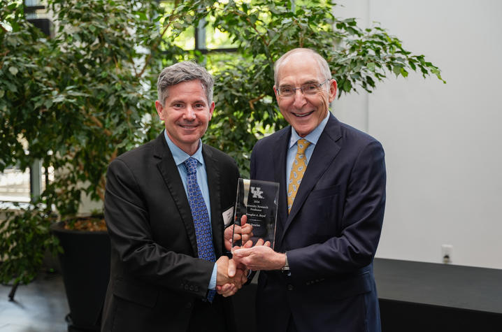 Two people in suits smile and shake hands while holding a glass award engraved “2026 University Research Professor.” They stand indoors with greenery and natural light visible behind them.