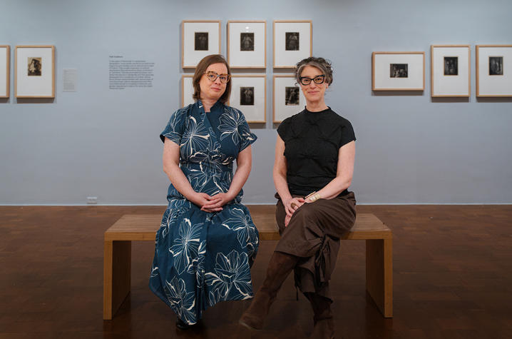 photo of Hooper and Schinberg Two people sit side by side on a wooden bench in a gallery space, facing the camera. Framed black-and-white photographs line the wall behind them in a neat arrangement, with soft lighting and polished wood floors creating a quiet, museum-like setting.