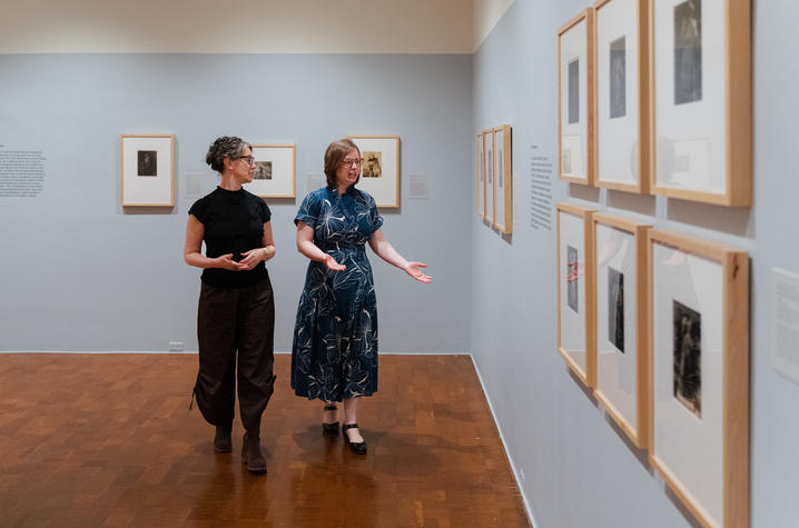 photo of exhibit Two people walk side by side through a gallery, looking at framed photographs hung in a row on a light-colored wall. The space is open and evenly lit, with wooden floors and descriptive wall text visible.