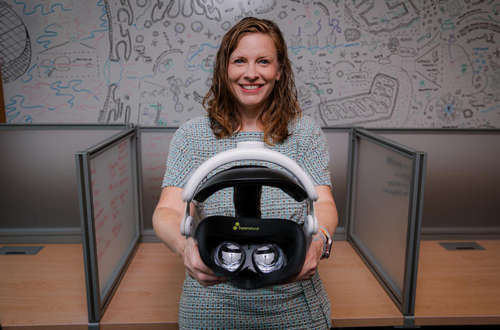 photo of Brandi Frisby with goggles Person standing between office cubicles holds a virtual reality headset toward the camera. A wall-sized whiteboard covered with colorful sketches, diagrams and notes fills the background of the workspace.