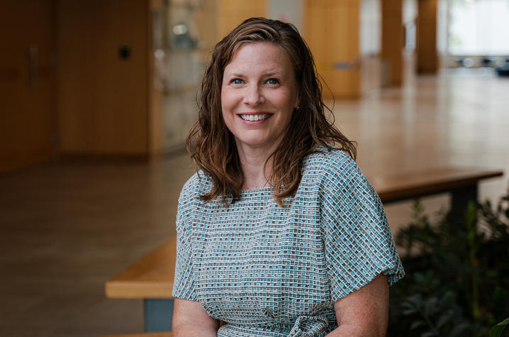 Brandi Frisby headshot Person seated on a wooden bench in a bright indoor hallway. Wooden walls, glass displays and plants line the space, with the corridor extending into the background.