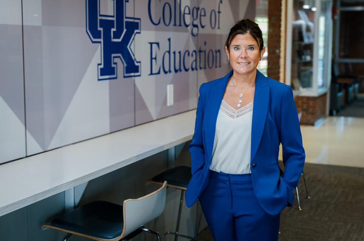 photo of Jena Hinds A person in a blue suit stands in a hallway beside a wall displaying the University of Kentucky logo and the words “College of Education.” A counter with stools lines the wall, and the hallway extends into the background.