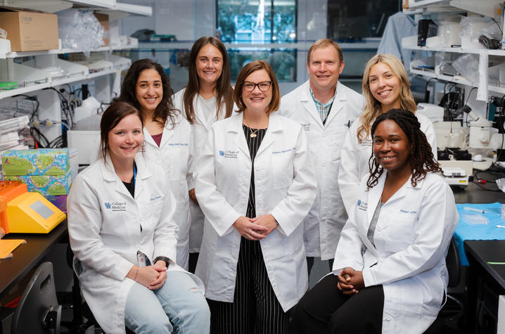 Macauley’s lab Research team members wearing lab coats pose together in a biomedical laboratory surrounded by microscopes, instruments and research supplies. The group photo highlights a collaborative team conducting laboratory research in neuroscience and medicine.