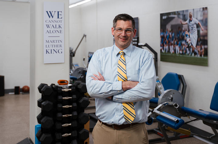Person stands with arms crossed in a biomechanics lab containing dumbbells, exercise equipment and wall displays, including a sign reading, “We cannot walk alone.”