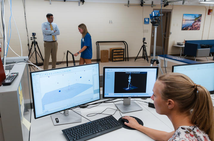 Person works at a computer displaying 3D motion-capture data while, in the background, another person walks through a biomechanics lab as an observer watches.