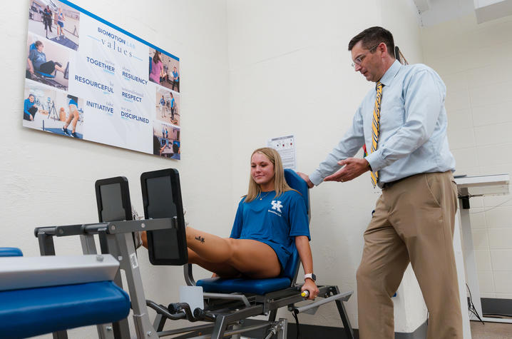 Person uses a seated leg exercise machine while another person stands nearby. A Biomechanics Lab values display hangs on the wall.
