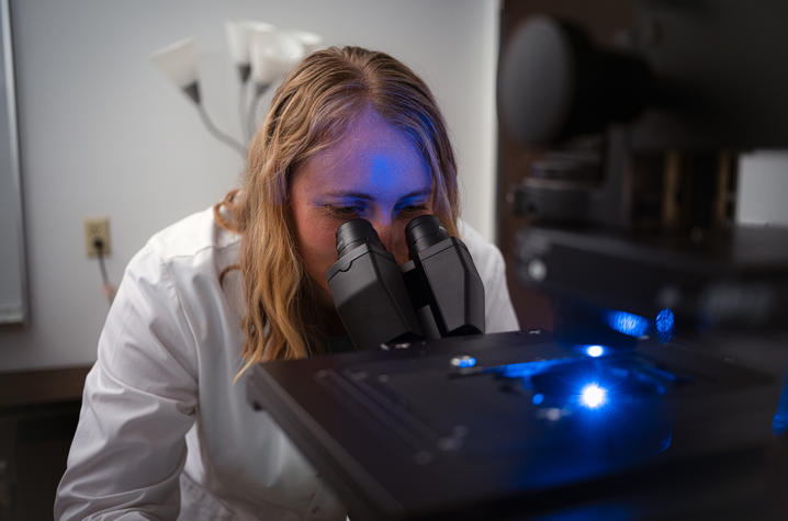 Long at microscope Person wearing a lab coat looks through a microscope in a research laboratory. Blue light from the instrument illuminates part of the workspace.