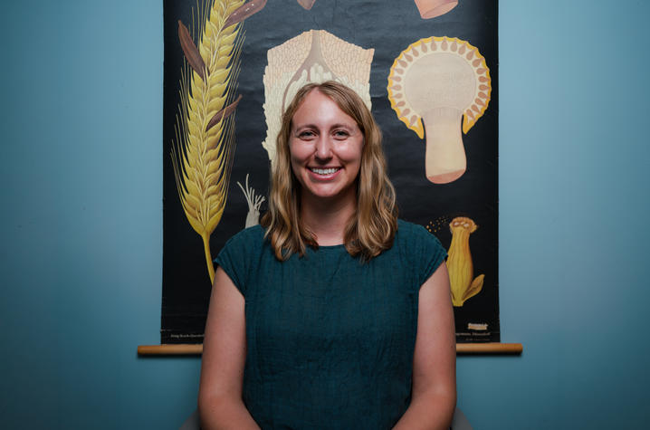 Long and illustrations Person seated in a chair poses in front of a botanical wall chart featuring enlarged illustrations of fungal structures on a dark background.