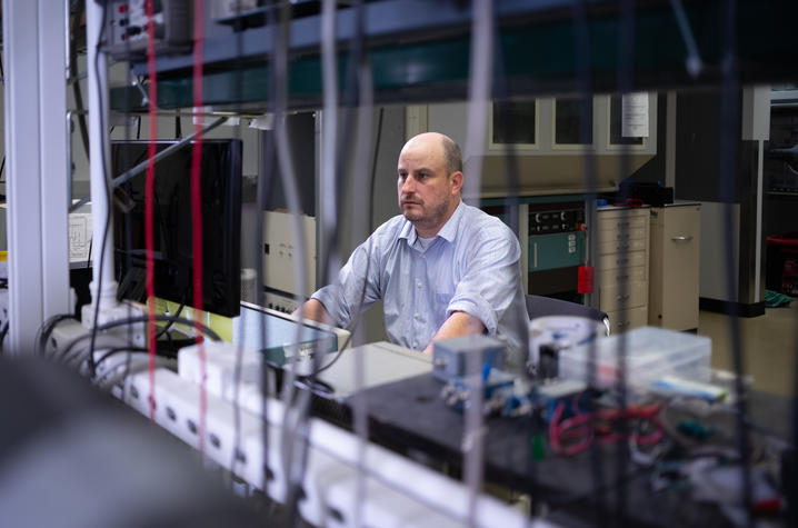 photo of William Gannon A researcher sits at a computer workstation in a laboratory, viewed through an array of cables, instruments and scientific equipment.