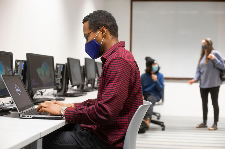 student in foreground on laptop at flex desk area on campus; students in background talking.