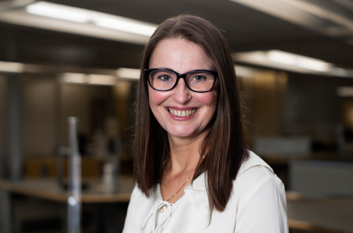 Close-up portrait of a person wearing glasses and a light blouse, looking at the camera in a softly lit indoor setting. The background is blurred, suggesting a modern interior with warm-toned lighting and structural lines overhead.