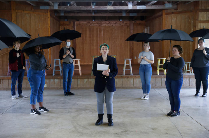 photo of cast members holding umbrellas surrounding other cast member reading from notepad in UK Theatre's "The Laramie Project"