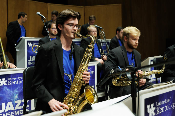 white male in a black jacket and blue shirt playing tenor saxophone in a jazz ensemble