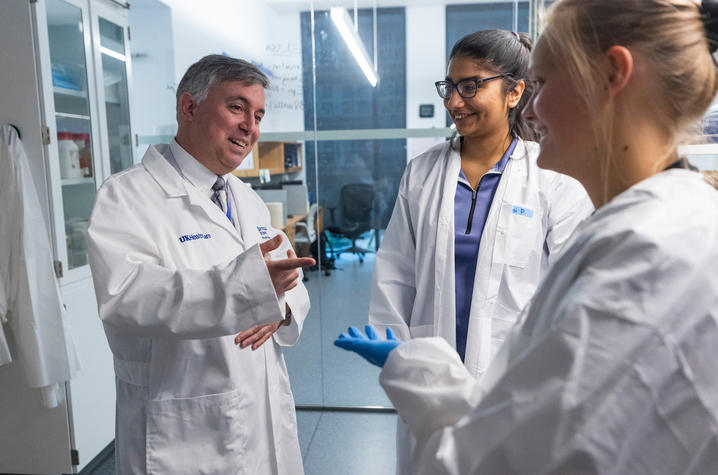 Three individuals wearing white lab coats are standing in a laboratory, engaged in discussion. One person is gesturing with a hand, while another holds a blue glove in hand. 