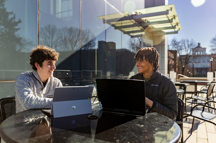 Two people sit outdoors at a table with laptops open, working or talking in a sunny area beside a glass building. Trees and additional buildings are visible in the background.
