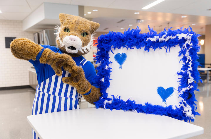  A costumed animal mascot wearing a blue-and-white striped apron stands behind a counter, holding a fluffy blue picture frame with a blank white center and a small blue heart, inside a bright cafeteria or kitchen setting.