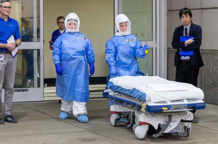 Two healthcare workers in full protective gear stand beside a hospital gurney outside a building, with two people in regular clothing nearby.