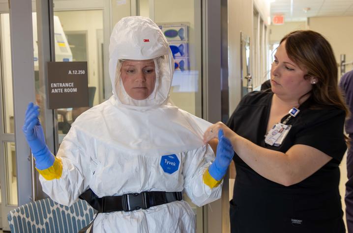 A person wearing protective coveralls and gloves stands in a hallway while another person adjusts the hood of the suit.