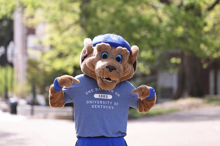 A University of Kentucky Wildcat mascot stands outdoors wearing a blue “One University at Kentucky” T-shirt.