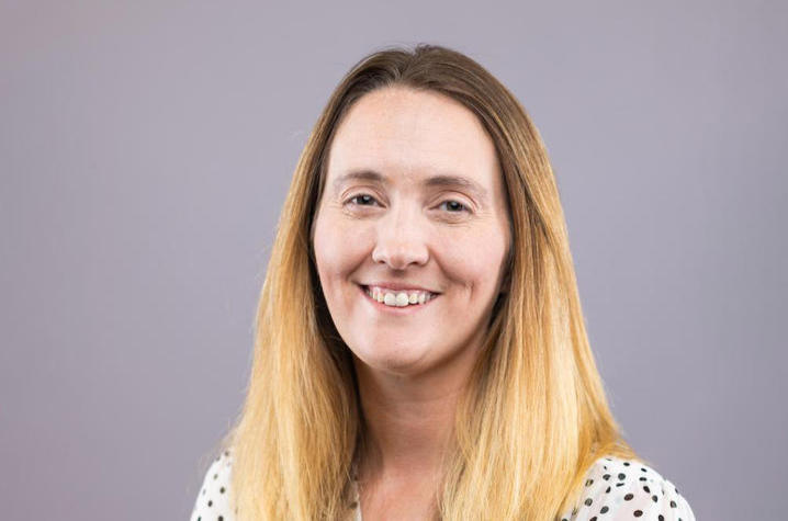 Portrait of a smiling woman with long blonde hair wearing a white polka-dot blouse against a light gray background.