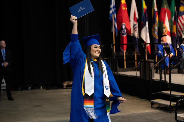 Graduate in blue cap and gown holding a diploma aloft on stage, wearing multiple stoles including a colorful striped one, with international flags displayed in the background.
