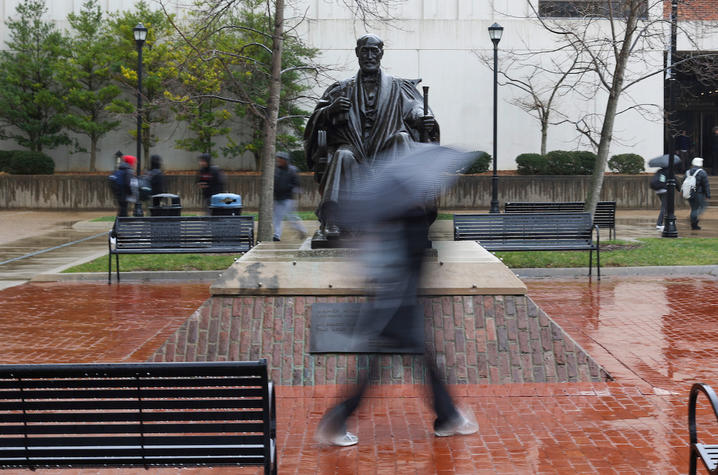 A photo of a student walking in front of the Patterson statue in the rain with a black umbrella.