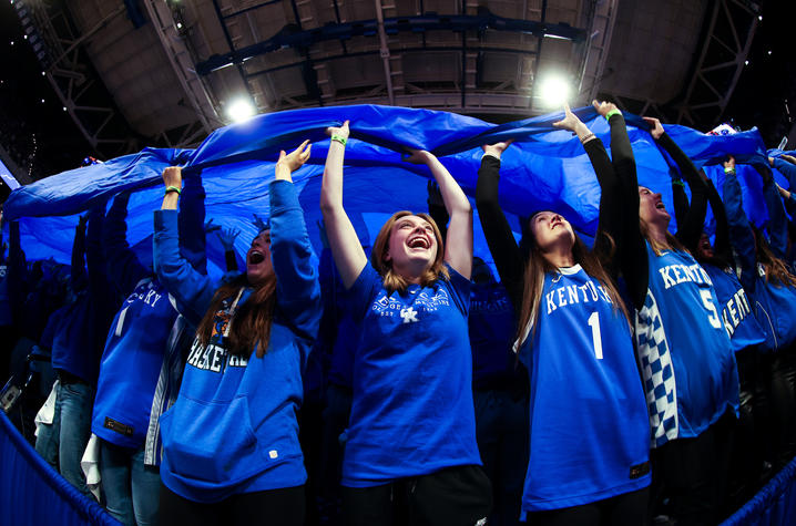 Fans in blue shirts hold a large blue banner overhead inside an arena.