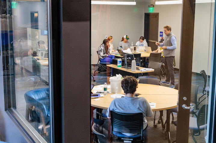 A view through a glass window into a classroom or meeting room where several people are seated around tables, engaged in discussion or work.